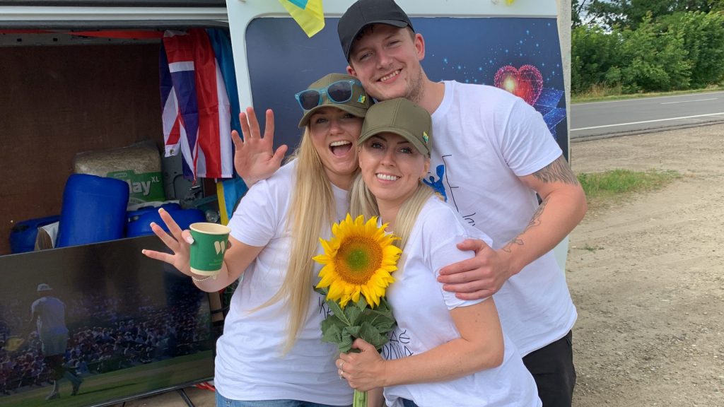 Three ABW volunteers in white t-shirts standing in front of a van used to deliver aid holding a sunflower in front of them