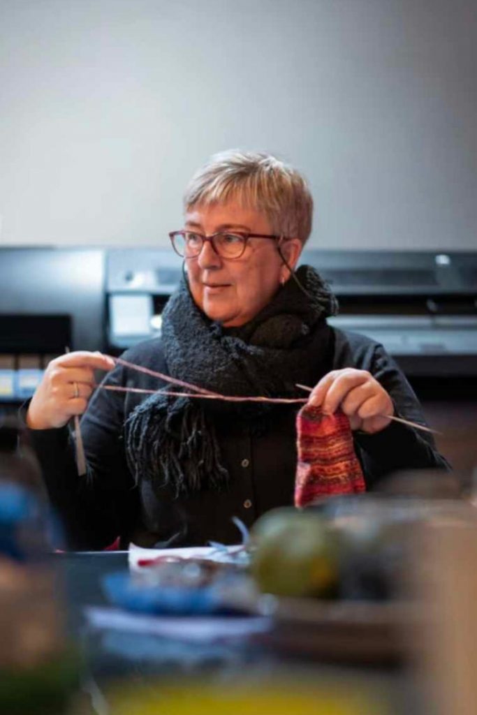 Berit from Paracrew in candid shot knitting warm woollen cloths to be sent into Ukraine from out base in Poland.  in her left hand white holding a thread of wool and a knitting needle in her right hand