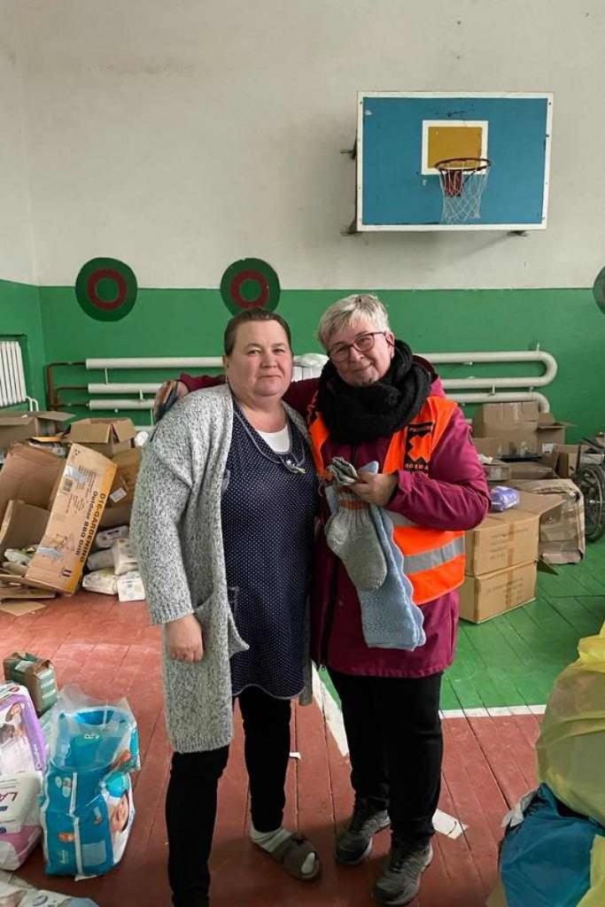 Berit from Paracrew, is in a gymnasium standing next to a Ukrainian woman. There are empty boxes scattered in the background where delivered aid has recently been unpacked. A basketball hoop with a blue backboard is mounted on the wall.