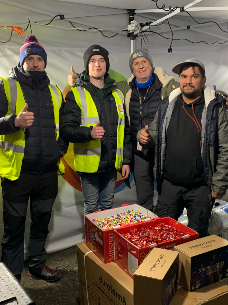 Four volunteers standing side by side in a tent with boxes of chocolates in front of them. They are preparing to distribute them to Ukrainian refugees fleeing the war.