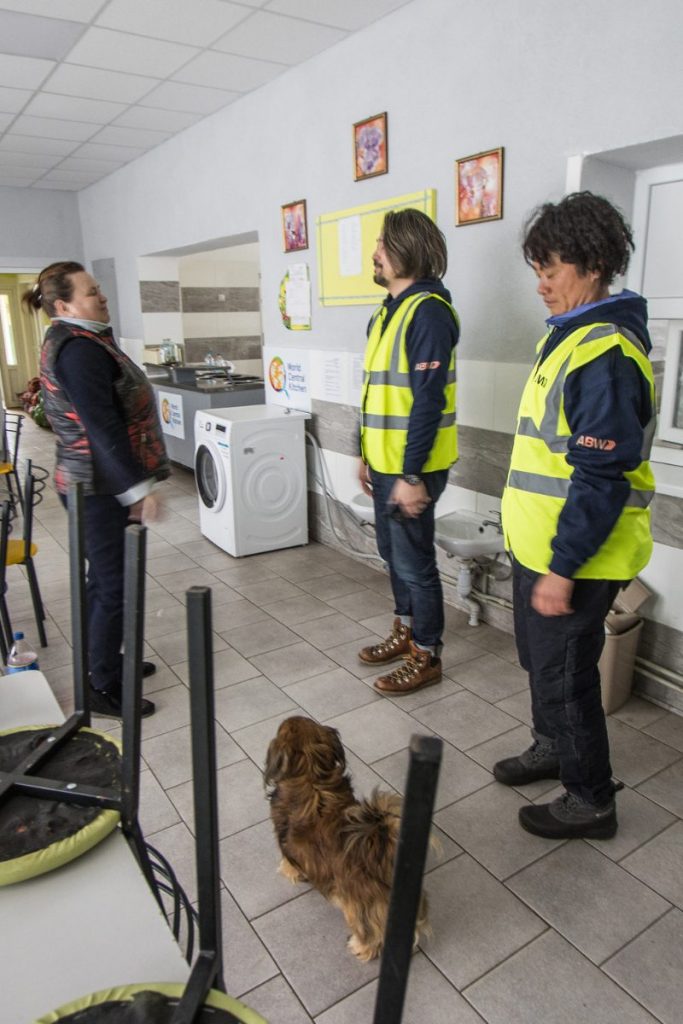WDRAC members Akira and Abe-Chan speaking to a local Ukrainian during an aid drop off inside of their facility. A small brown dog is seen in the bottom of the frame and Abe-Chan is seen glancing down at it.